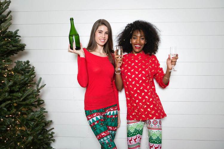 Two women in holiday clothes holding champagne glasses and smiling by a Christmas tree.